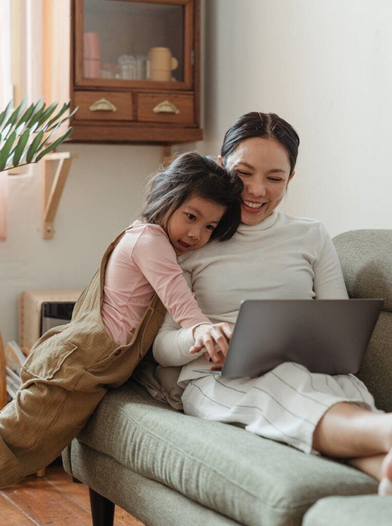 Cheerful smiling Asian woman browsing modern netbook while hugging with cute content daughter on comfy sofa in cozy living room emphasizing the importance of Teaching Kids About Money: Financial Literacy For Children