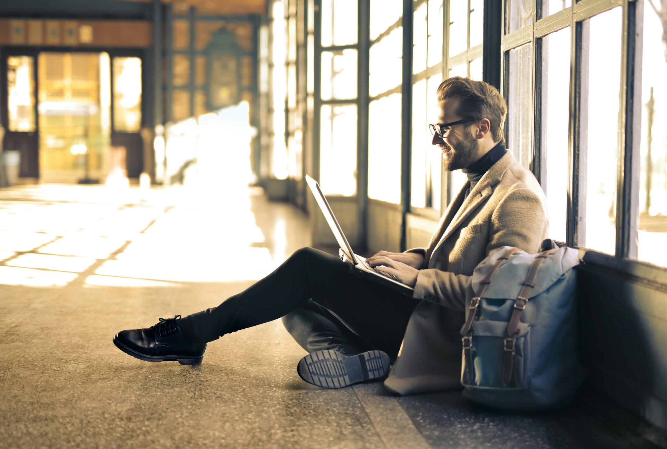 a man in his 40s dressed nicely and sitting on the ground in what looks like an open loft workspace with his back against a wall full of mirrors. he's looking at his laptop on his lap with a big smile on his face, capturing the joy of it not being too late for investing for retirement in your 40s