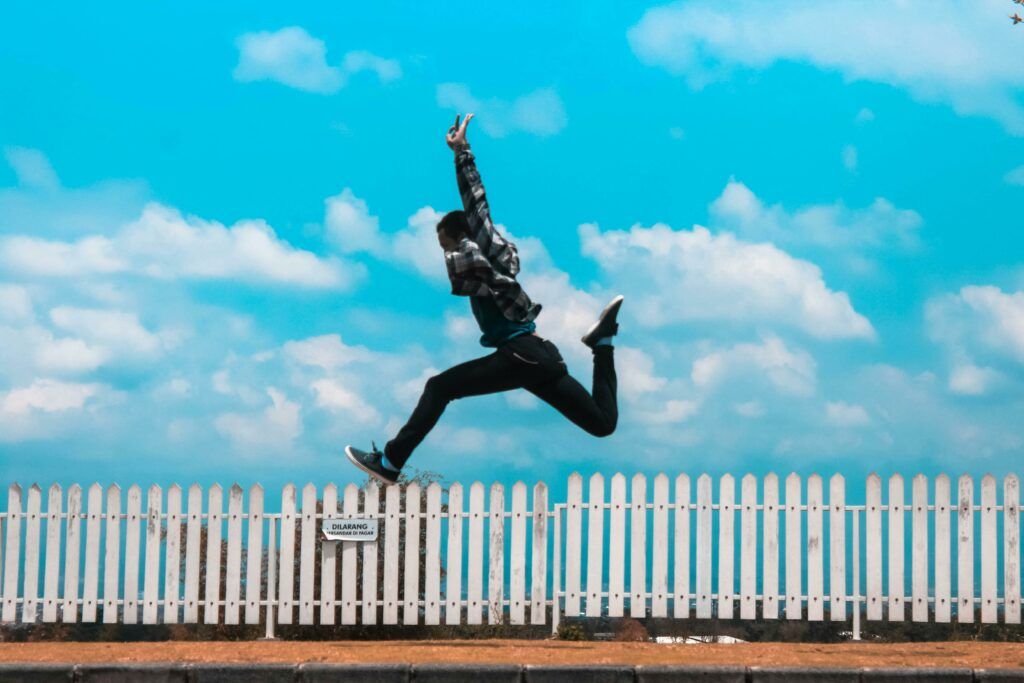 side vie of a person jumping in a running position with arms straight up in front of a white picket fence and blue skies and puffy white clouds in the background, symbolizing the joy of breaking through.