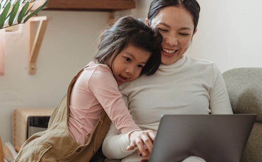 Cheerful smiling Asian woman browsing modern netbook while hugging with cute content daughter on comfy sofa in cozy living room emphasizing the importance of Teaching Kids About Money: Financial Literacy For Children