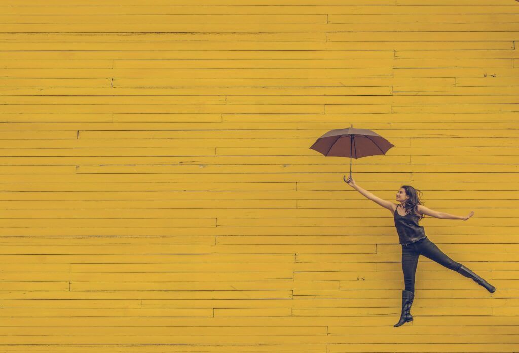 A woman holding brown umbrella in front of a yellow wall that completely fills the image, looking like she's floating upward, reflecting the idea of personal development ang going higher and higher